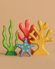 The Handmade Holzwald Coral Reef Set features colorful wooden toys shaped like seaweed, coral, and a blue starfish with white spots, all arranged against a plain beige background.