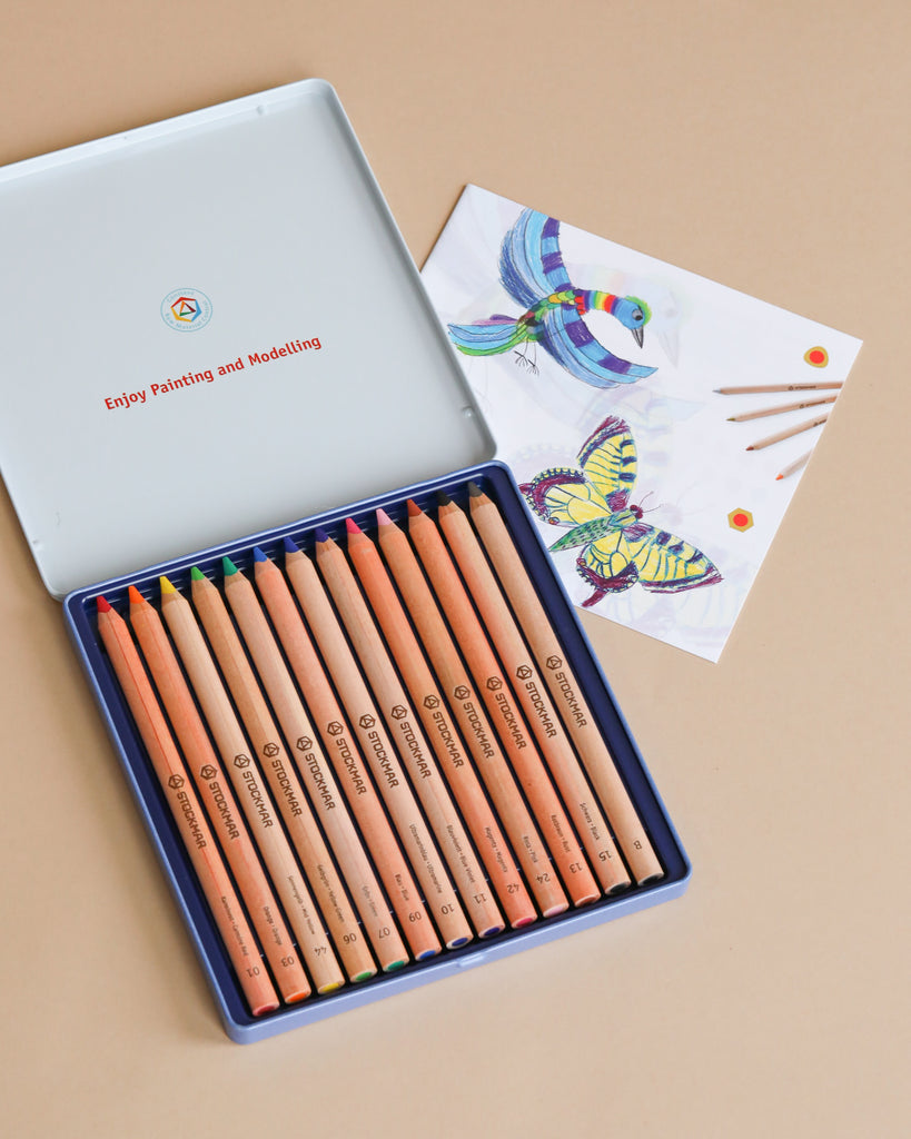 An open Easter Basket Gift Set displays 12 colored pencils arranged neatly in a tin box, next to a Kids Drawing Book with a bird and butterfly illustration, plus three pencils on a beige background.