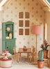 The Maileg Fully Furnished Dollhouse Set + Surprise Gift includes a charming dining room scene with a table, four chairs, green cabinet with dishes, pink polka-dot lamp, cakes on display, potted plants, and patterned wallpaper.