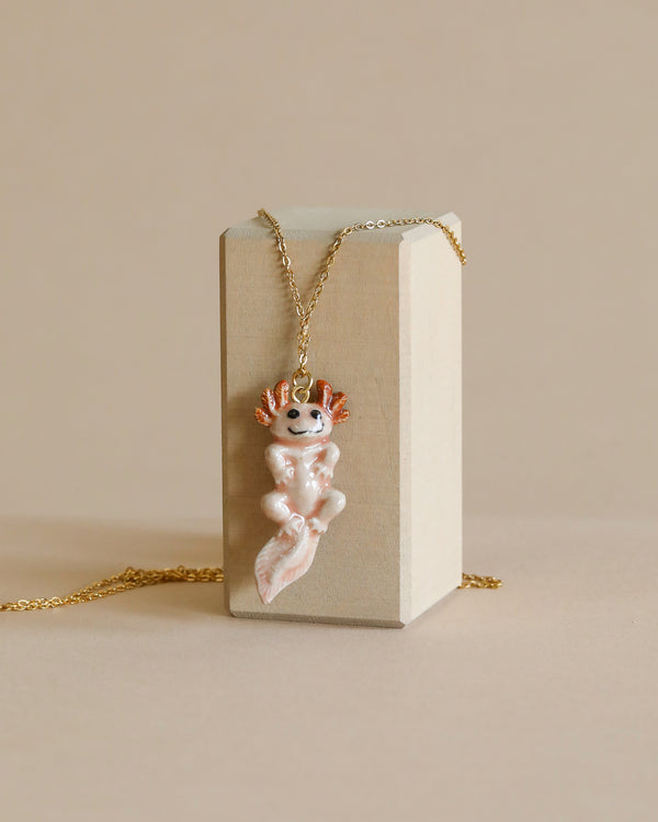 A whimsical necklace featuring a handcrafted porcelain Axolotl Salamander pendant with smiling facial features hangs on a thin 24k gold plated chain draped over a rectangular beige block against a light