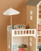The Maileg Fully Furnished Dollhouse Set + Surprise Gift features a balcony with charming furniture—a white table, two chairs, a green watering can, small potted plants, and a white umbrella—attached above the detailed dollhouse rooms.