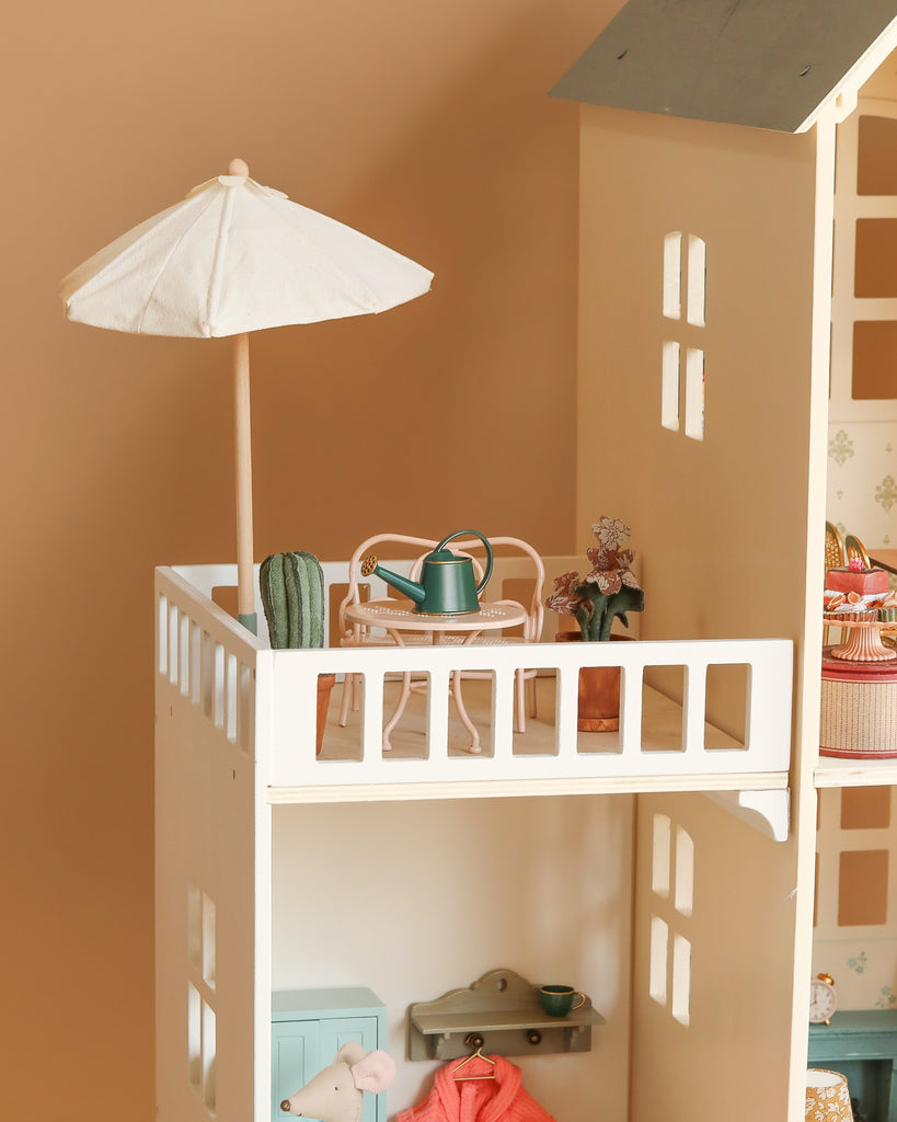 The Maileg Fully Furnished Dollhouse Set + Surprise Gift features a balcony with charming furniture—a white table, two chairs, a green watering can, small potted plants, and a white umbrella—attached above the detailed dollhouse rooms.