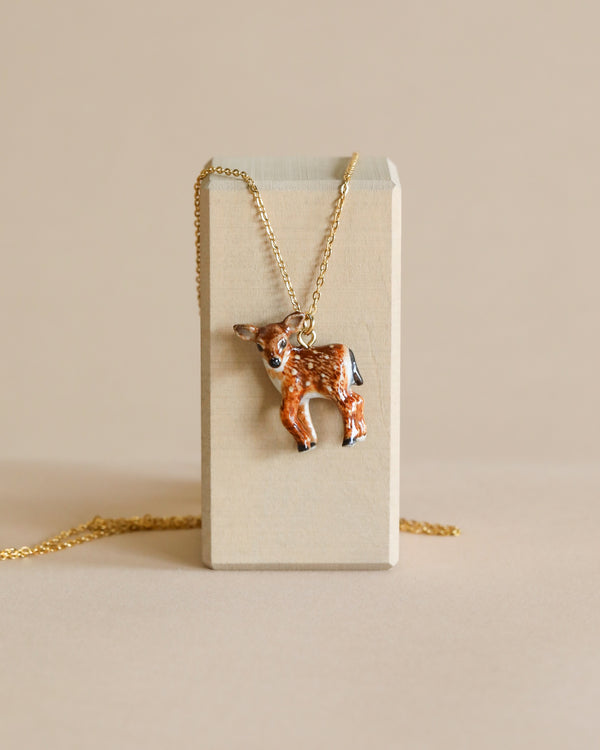 A Fawn Necklace, hand painted and hanging on a 24k gold plated chain, displayed against a beige block on a light background.