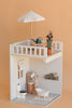 A miniature dollhouse room displays the Maileg Bonus Room Bathroom Set with a bathtub, toilet, vanity, and shelf. On the rooftop patio are mini accessories—a table, chairs, umbrella, and potted plants—set against a peach background.