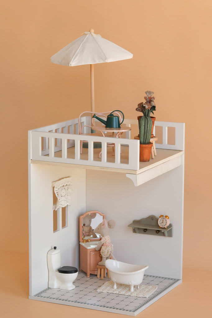 A miniature dollhouse room displays the Maileg Bonus Room Bathroom Set with a bathtub, toilet, vanity, and shelf. On the rooftop patio are mini accessories—a table, chairs, umbrella, and potted plants—set against a peach background.