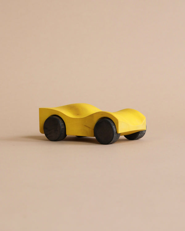 A simple, yellow Handmade Wooden Race Car - Yellow made from beech with black wheels is shown against a plain beige background. The car, painted with non-toxic paint, has a sleek, minimalist design with smooth curves and no additional details.