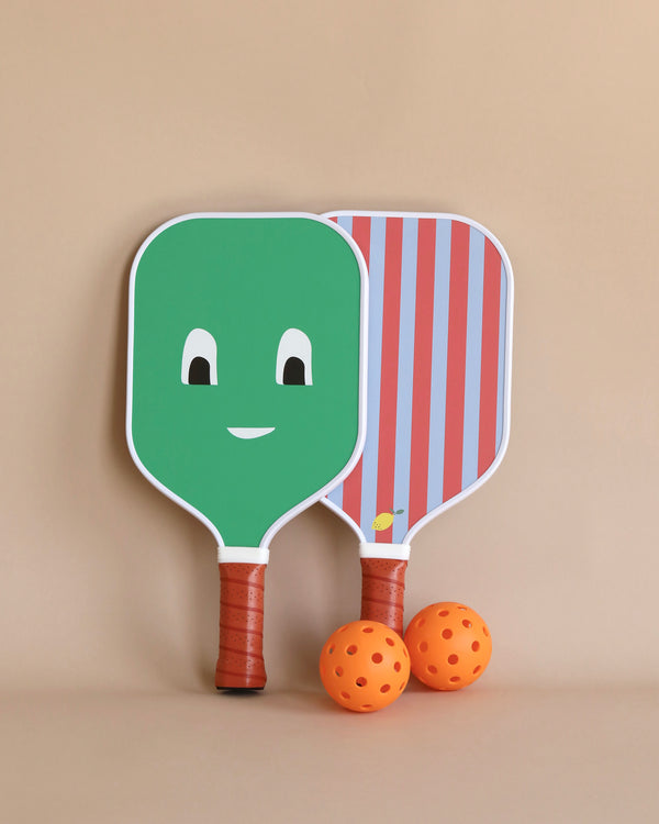 Two pickleball paddles with cartoon faces and two orange perforated balls against a beige background. One paddle, measuring 7.4 x 15 inches, is green with a smiling face, while the other in the Sticky Lemon Pickle Ball Game Set has a striped red and blue face featuring an image of a pickleball.