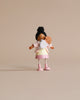 A small doll with dark hair styled in a bun, wearing a white and pink dress made of knitted and jersey fabrics, holding Mrs. Forrester and the Baby against a plain beige background.