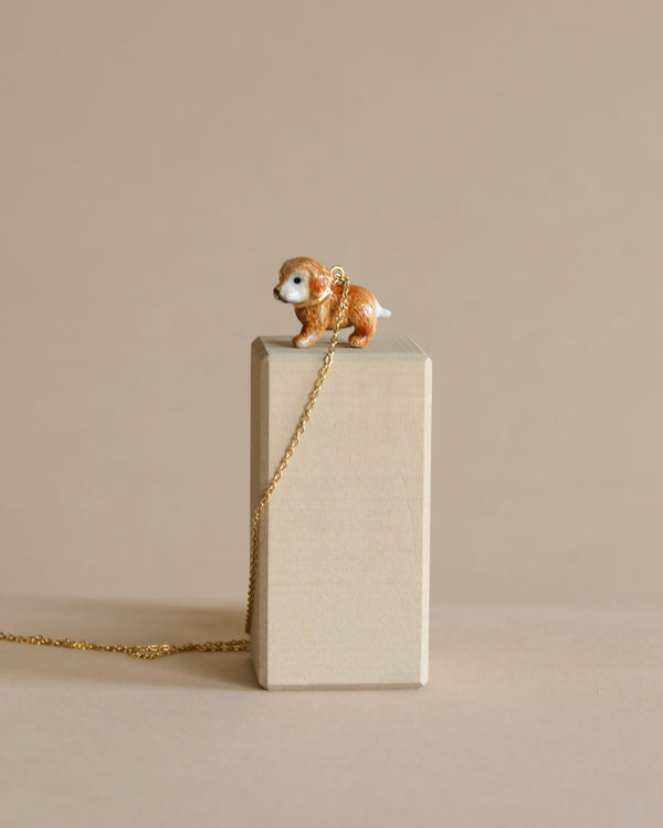A small figurine of a Golden Retriever Necklace perched atop a beige, rectangular block, with a 24k gold plated chain draped beside it on a neutral background.