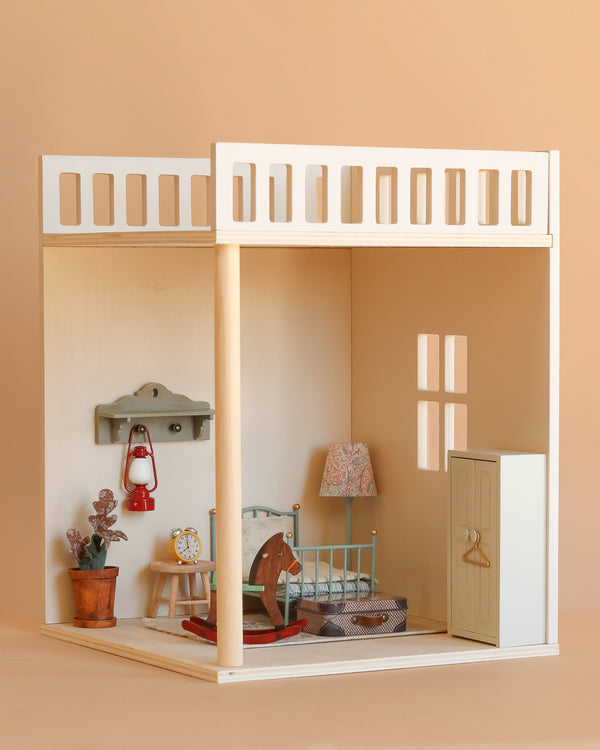 The Maileg Bonus Room Bedroom Set features a bed, rocking horse, lamp, plant, clock, red lantern shelf, and wardrobe—all arranged in a cozy miniature room with beige walls, a window, and charming dollhouse accessories.