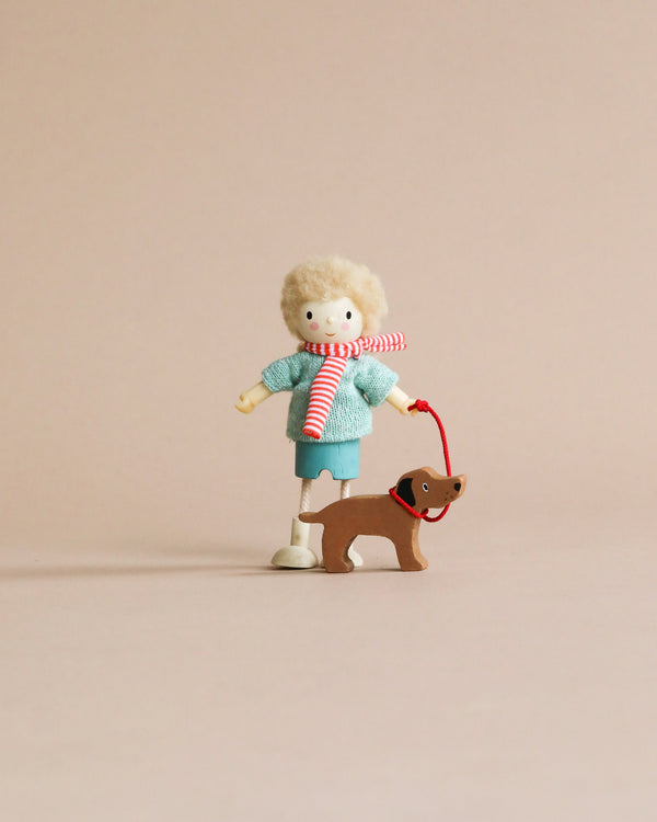 A small doll with curly hair and a blue sweater holds a red and white striped leash attached to Mr. Goodwood and his Dog, on a plain beige background.