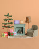 The Maileg Christmas Morning Living Room Set includes a fireplace, decorated Christmas tree, wrapped presents, floral lamp, dollhouse accessories, checkered armchair, and patterned rug arranged on a beige background.