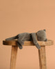 The Maileg Sleeping Kitten Plush, Medium - Grey, made from recycled polyester, is lying on its side on a wooden stool against a plain beige background.