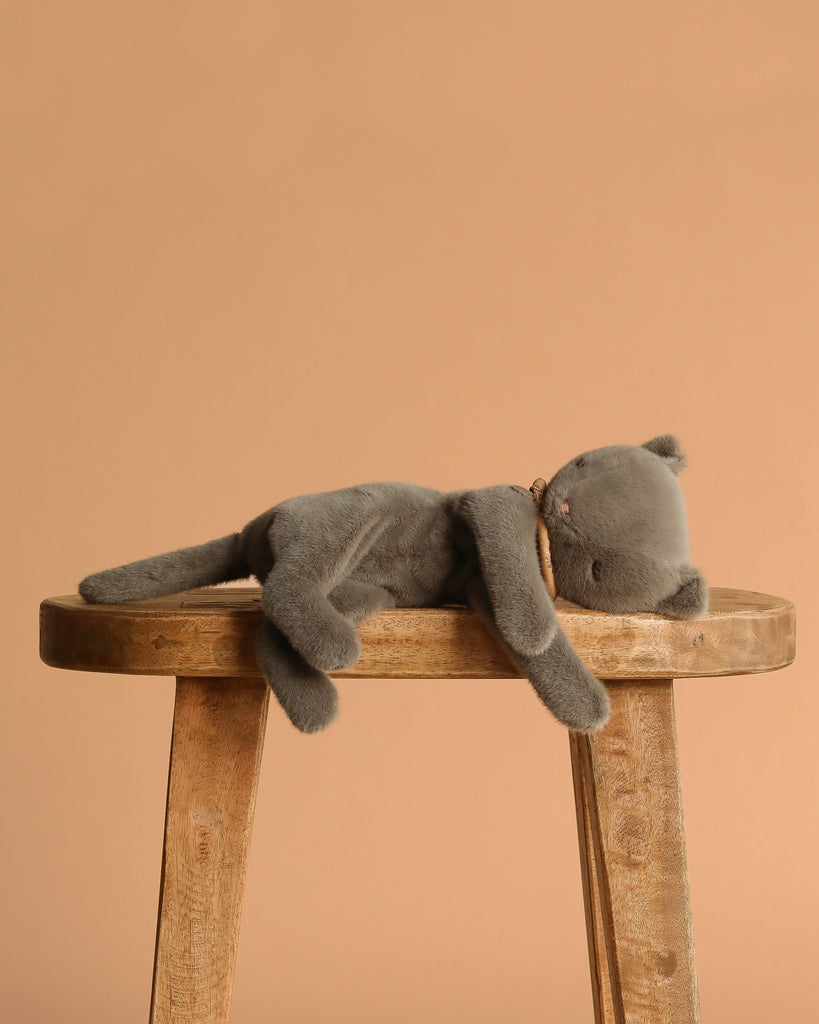 The Maileg Sleeping Kitten in Carry Cot, a gray plush kitten, lies stretched out on its side on a wooden stool against a plain beige background.