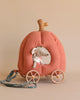 A small stuffed mouse toy from the royal mice family sits inside a soft, fabric fairytale carriage with a handle for pulling. The Maileg Pumpkin Carriage is orange with a stem on top and four wheels at the bottom. The mouse, dressed in a white outfit, peeks through a window in the carriage.