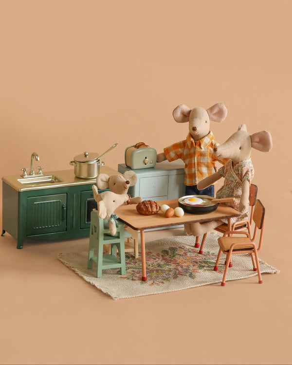 Three Maileg Mice enjoy a cozy Maileg Kitchen scene with the Maileg Family Breakfast Set, featuring a stove, sink, table, and food items. One mouse sits at the table while another stands nearby and an adult mouse cooks breakfast.