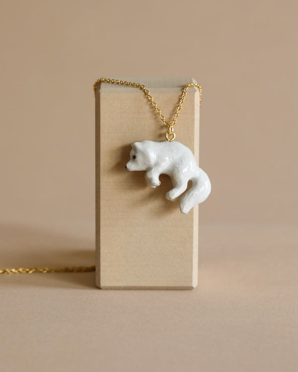 A fine porcelain Polar Fox Necklace hanging on a 24k gold plated chain draped over a vertical tan card holder set against a neutral beige background.