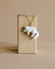 A fine porcelain Polar Fox Necklace hanging on a 24k gold plated chain draped over a vertical tan card holder set against a neutral beige background.
