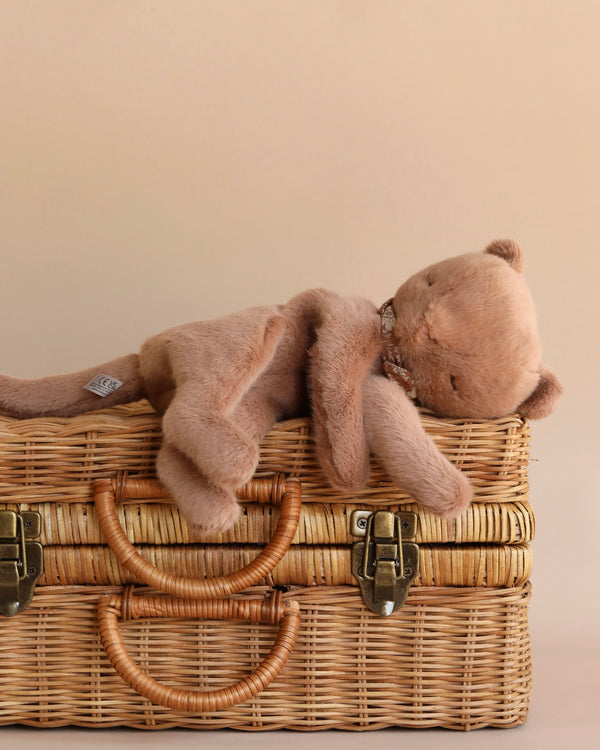 A Maileg Plush Kitten, Nougat lies on its stomach atop a cosy wicker picnic basket, with its limbs draped over the edges. The background is a warm beige color, complementing the tones of the kitten and the basket.