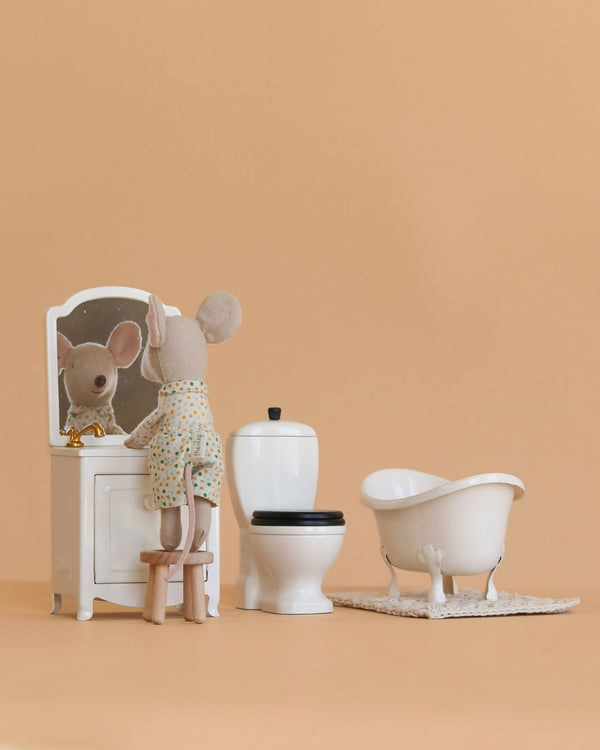 A plush mouse in pajamas stands on a stool, gazing into a vanity mirror beside the Maileg Little Bother Bathroom Set and Maileg furniture, all set against a peach backdrop inspired by the Mouse Hole Farmhouse.