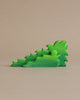 A vibrant green, foam dinosaur puzzle piece, coated with non-toxic paint, stands upright against a plain beige background. The piece is detailed with various spikes and curves, resembling a stegosaurus