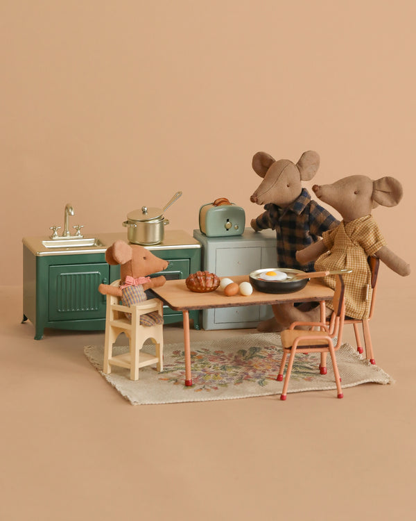 Three toy mice, including a Maileg Mouse, gather around a kitchen table with food, while the Maileg Family Breakfast Set—with stove, sink, and accessories—is displayed in the background on a beige backdrop.