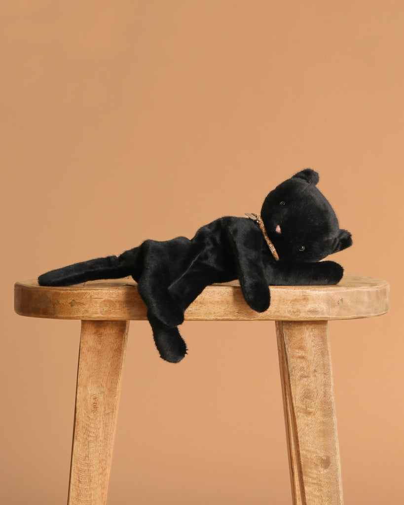 The Maileg Sleeping Kitten Plush, Medium - Black, with a beige ribbon collar, is lying on its side on a wooden stool against a plain light brown background.