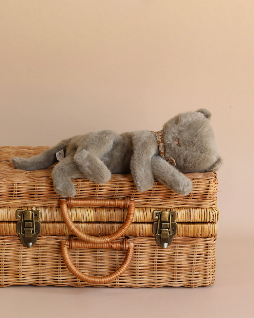 A Maileg Plush Kitten, Earth grey lies on its side atop a cosy, woven picnic basket with tan leather handles and brass latches. The background is a soft beige color, giving the scene a minimalistic and warm feel.