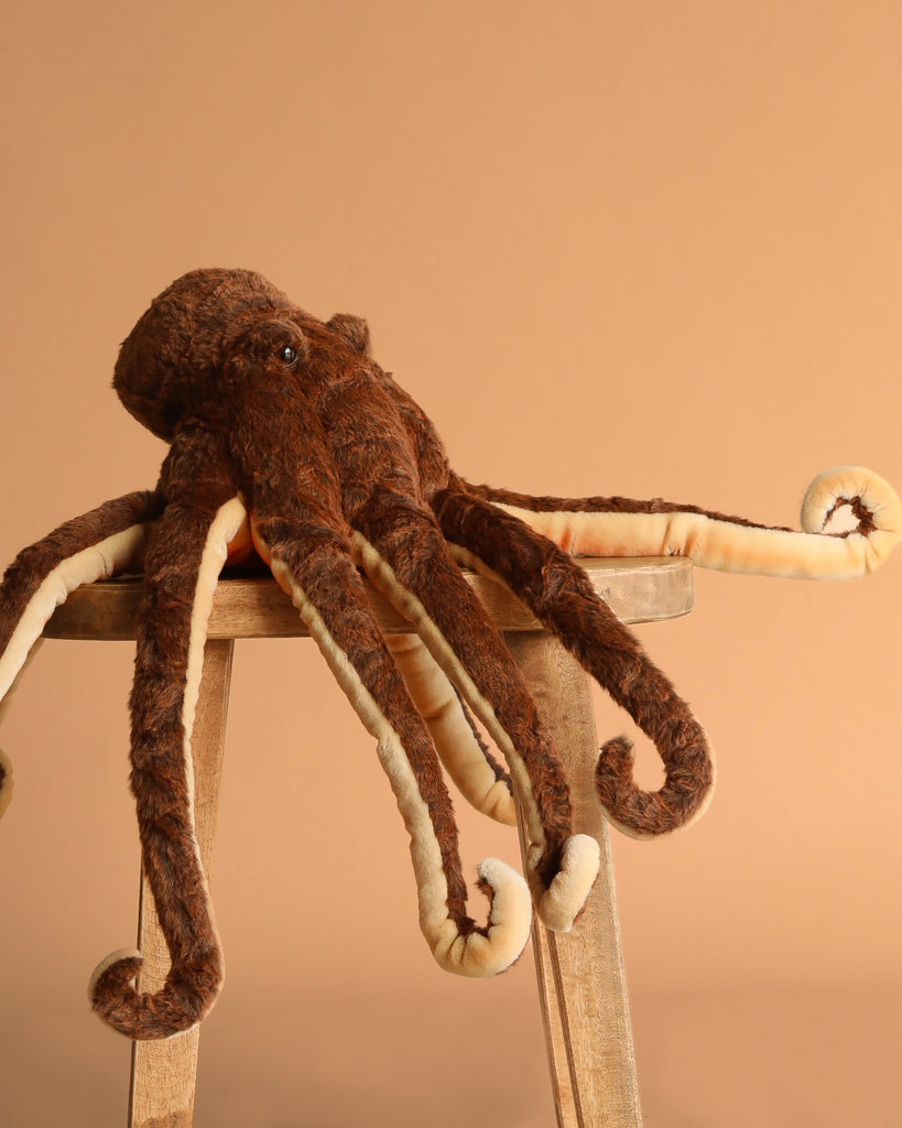 The Octopus Stuffed Animal features soft, brown plush with cream undersides and hand-sewn, curled tentacles. It sits draped over a wooden stool against a beige background, highlighting its realistic charm.