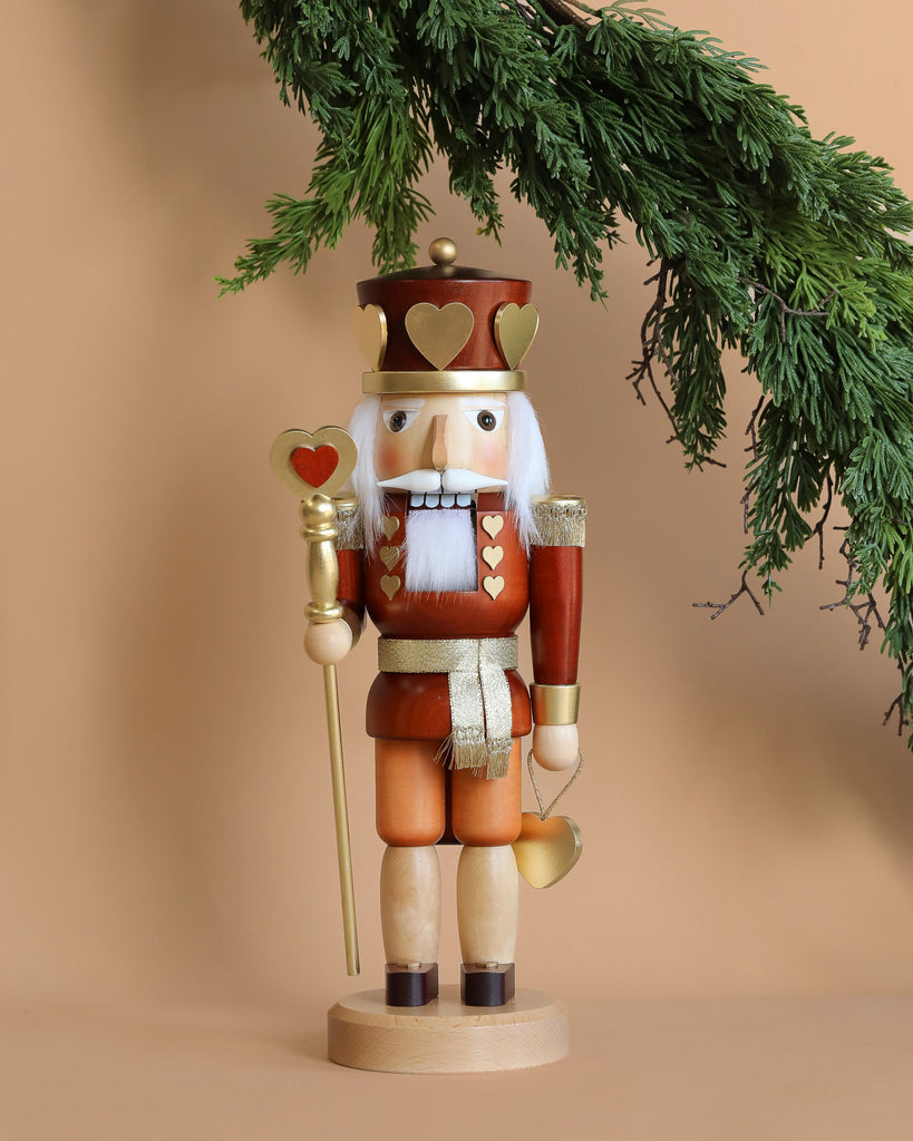 The Christian Ulbricht Collectible Wooden Nutcracker - King of Hearts, a limited-edition piece, stands proudly under a green pine branch against a beige background. Adorned with golden hearts, a crown, and a scepter, this meticulously crafted German creation exudes timeless charm.