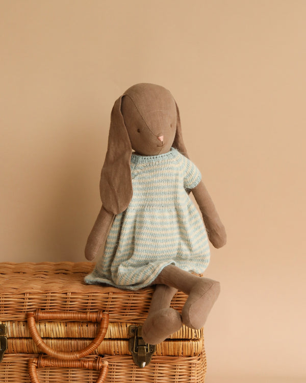 A soft, brown Maileg Bunny Size 4, Brown - Knitted Dress is seated on top of a closed wicker basket. Crafted from natural linen fabric, the bunny features long, floppy ears and a minimalist design. With its neutral beige backdrop, this charming companion is recommended for all ages.