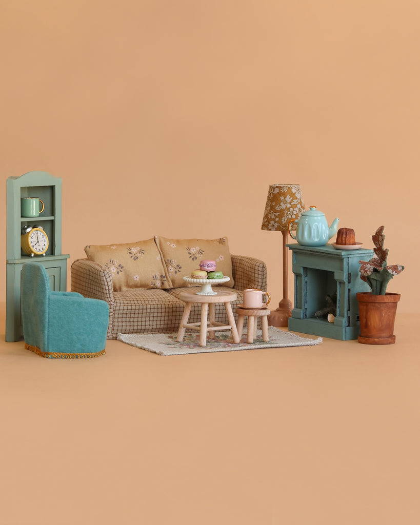 A charming miniature scene displays the Maileg Living Room Set, featuring a couch, armchair, side table with pastries, rug, potted plant, shelf with clock and lamp, and a blue fireplace with a tea set—all on a plain beige background.