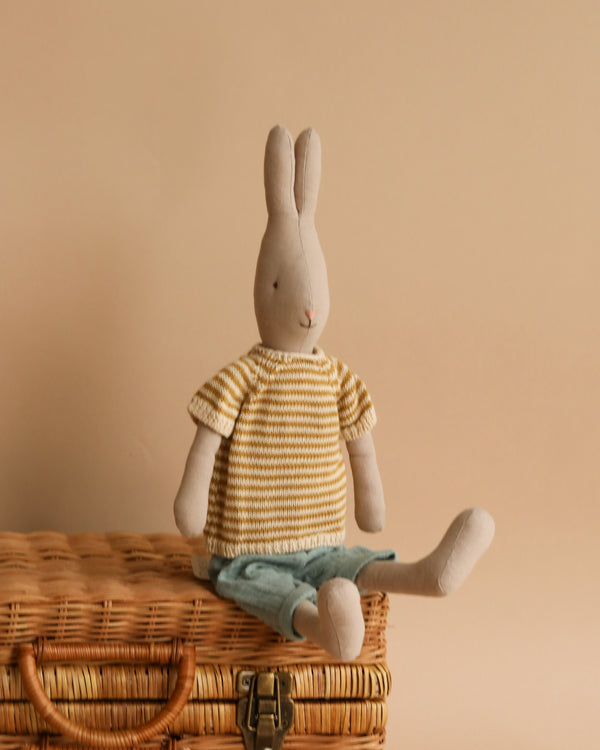 A Maileg Rabbit Size 3, Classic - Knitted Shirt and Pants with long ears sits on a wicker basket, its yellow and white striped sweater and blue pants making it a charming addition to any soft natural fabrics collection. The background is a warm beige color.
