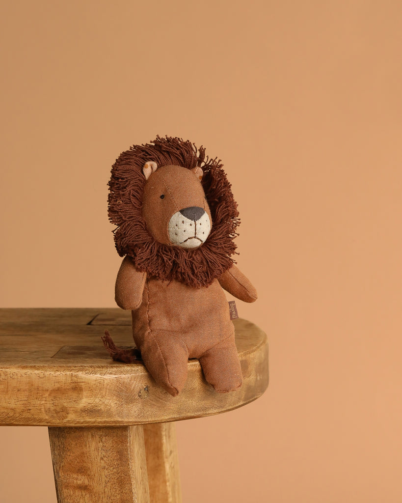 The Maileg Mini Safari Friends Box Set features a plush lion with a brown mane, shown sitting on a wooden stool against a light brown background—ideal for soft stuffed animal fans.