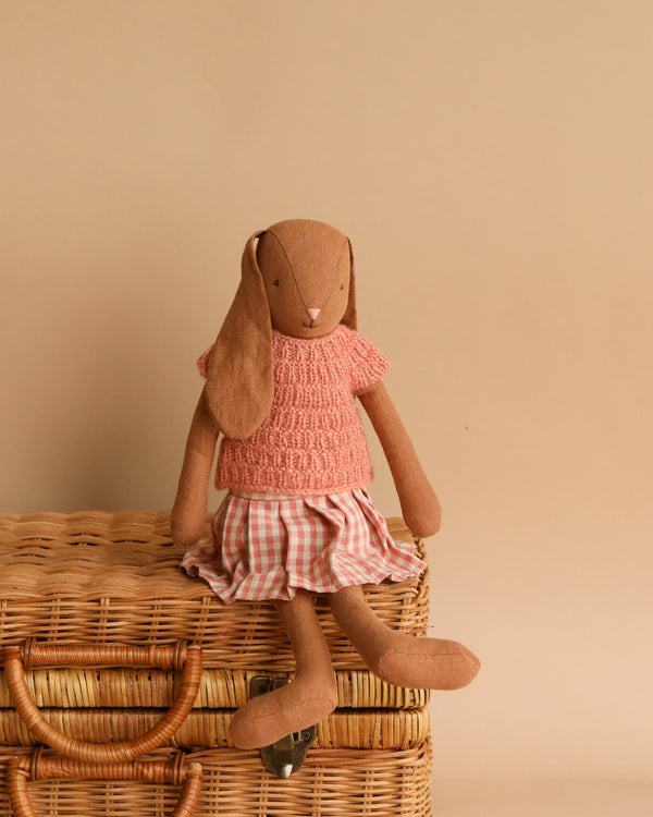 A Maileg Bunny, Size 3 - Knitted Shirt & Skirt sits on a woven wicker basket. The bunny is wearing a pink knitted top and a plaid skirt, all crafted from soft natural fabrics. The background is a plain beige color, enhancing the scene's cozy, eco-friendly feel with its use of recycled materials.
