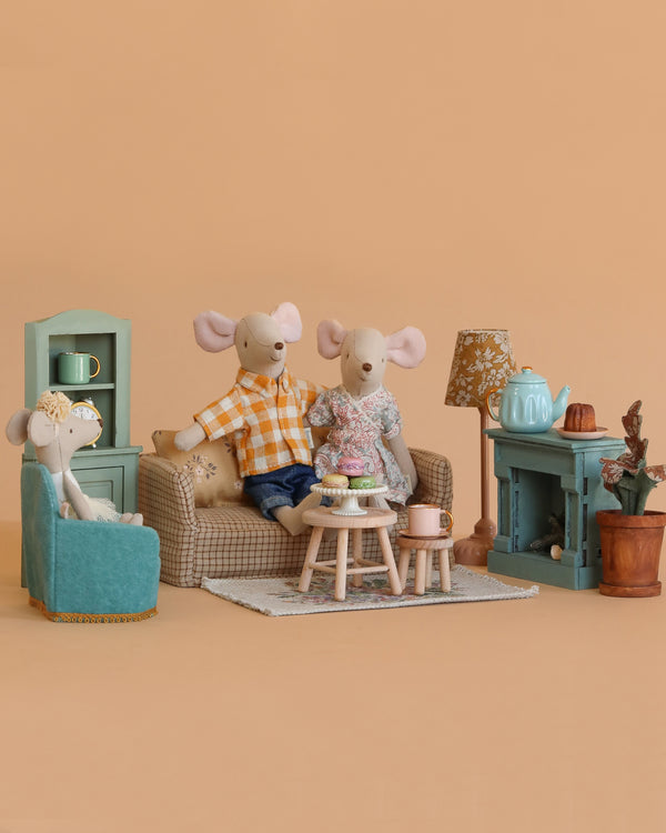 Three toy mice enjoy the Maileg Family Time Living Room Set, featuring mouse dollhouse furniture, a tea set, teal shelves, and a potted plant—all arranged in a cozy scene against a plain peach background.