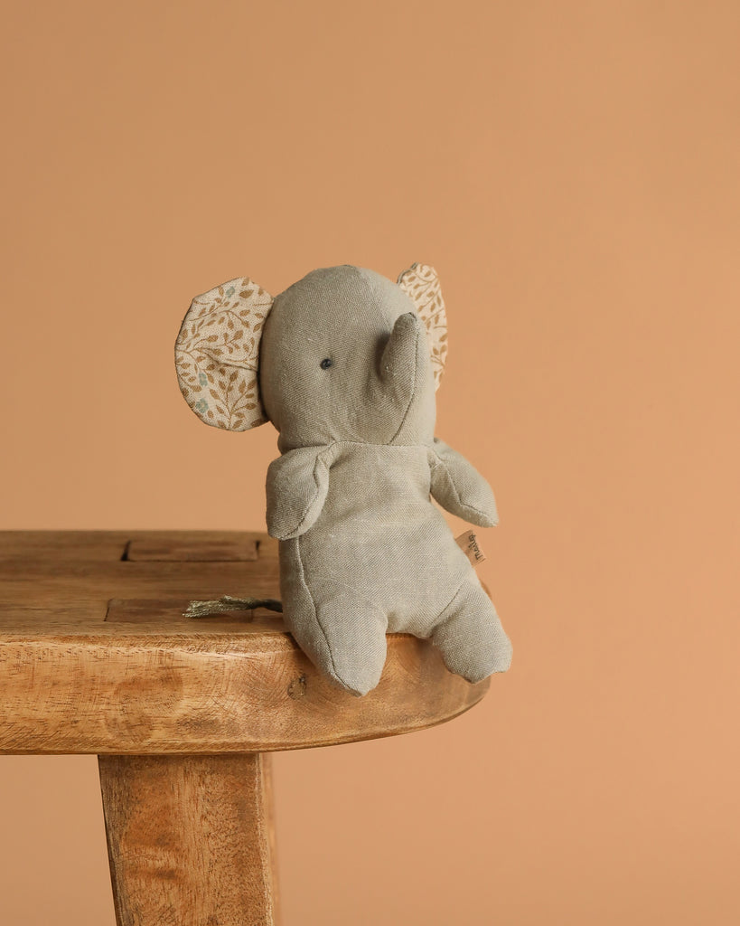 A small, gray elephant with patterned ears from the Maileg Mini Safari Friends Box Set sits on a wooden stool against a plain, light brown background—a charming addition to your soft animal collection.