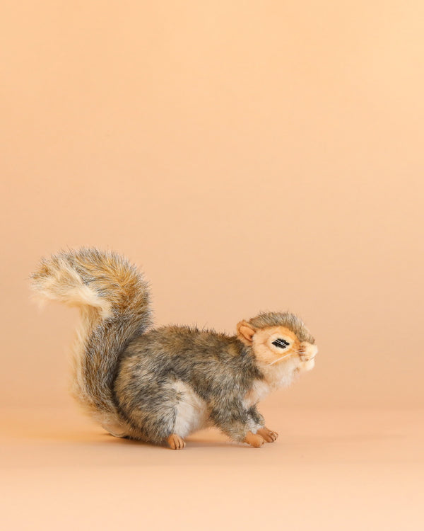 A Gray Squirrel Stuffed Animal with soft gray and white fur stands on a light peach background, facing right with its bushy tail raised; this charming plush toy is carefully hand sewn to highlight its lifelike features.