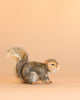 A Gray Squirrel Stuffed Animal with soft gray and white fur stands on a light peach background, facing right with its bushy tail raised; this charming plush toy is carefully hand sewn to highlight its lifelike features.