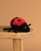 The Lady Bug Stuffed Animal features a red body with black spots and legs, and is displayed on a wooden stool against a plain brown background.