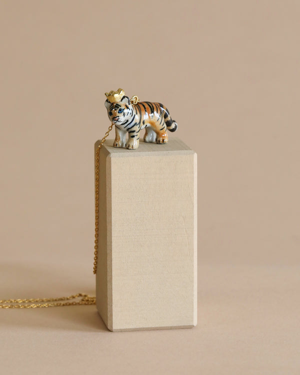 A small ceramic Tiger King Necklace figurine with a gold crown, perched on top of a rectangular beige pedestal, with a 24k gold plated chain necklace draped over it and trailing off to the side.