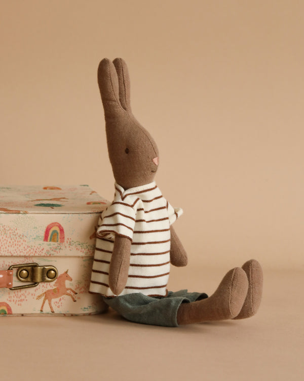 A Maileg Rabbit Size 2, Brown - Striped Blouse and Pants, made from soft natural fabrics, sits next to a decorative box with a latch. The backdrop is a solid beige color, and the box features illustrations of rainbows and deer.