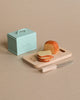 A wooden cutting board with a loaf of bread and slices on it, along with a small serrated knife, sits on a beige surface. Next to it is a light blue box with a handle and the word "Maileg Miniature Bread Box" embossed on the front, perfect for adding charm to your miniature kitchen.