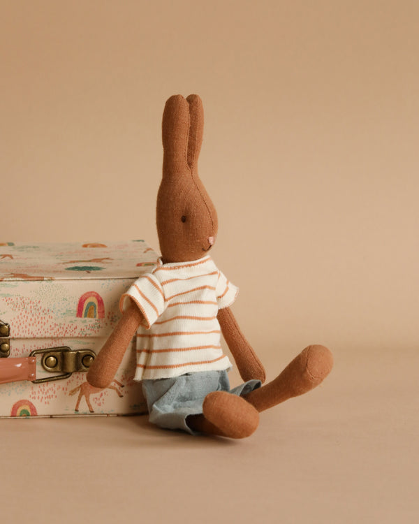 A soft brown stuffed bunny with long ears, clad in a white and orange striped shirt and denim shorts, sits next to a small, pastel-colored suitcase decorated with rainbows and plants. Made from soft natural fabrics, this Maileg Rabbit Size 1, Chocolate Brown - Striped Blouse and Shorts rests on a plain, light brown surface.