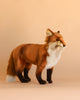 The Standing Red Fox Stuffed Animal, featuring brown fur, black legs, and a white chest, stands on a light beige background. Expertly artisan handcrafted with lifelike details and exquisite quality, this plush fox makes an ideal addition for collectors who appreciate realistic stuffed animals.