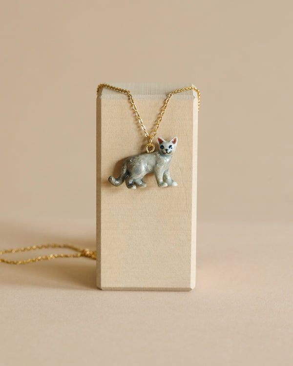 A Grey Cat Necklace with a gemstone eye is displayed on a small rectangular beige stand, with a 24k gold plated chain draped over it, set against a plain beige background.