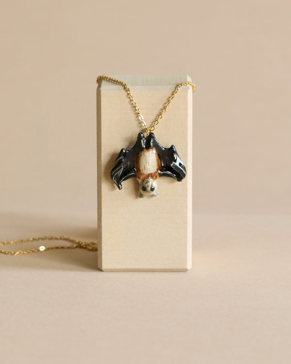 A black Fruit Bat Necklace with small hand-painted white details hanging on a gold chain displayed on a beige box against a matching beige background.