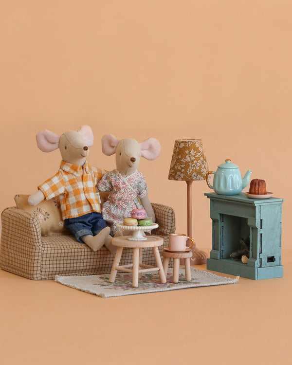 Two toy mice relax in the Maileg Mom and Dad Living Room Set, featuring a cozy couch, small table with pastries, tea set, lamp, blue fireplace, cups, and a warm beige background.