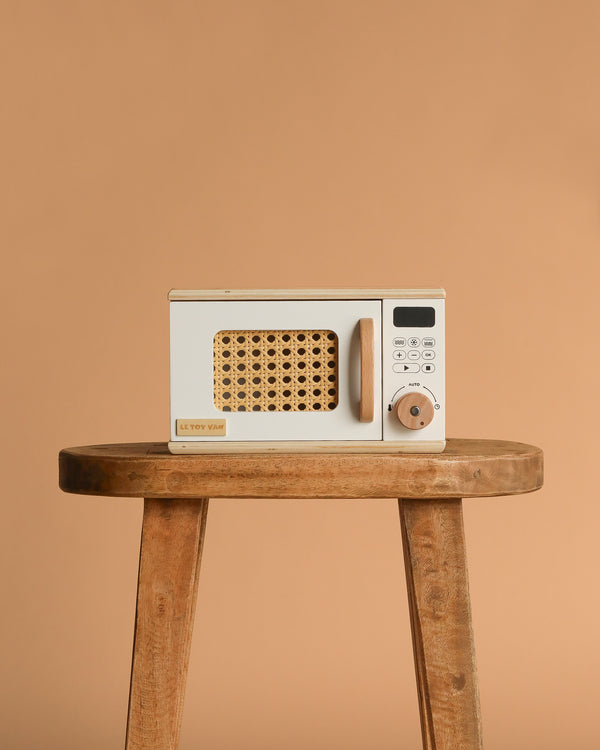 The Wooden Microwave, a small beige toy with a wicker-patterned door, sits on a sustainable wooden stool against a plain tan background—ideal for any play kitchen.