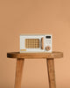 The Wooden Microwave, a small beige toy with a wicker-patterned door, sits on a sustainable wooden stool against a plain tan background—ideal for any play kitchen.
